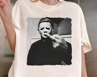 May include: Cream-colored t-shirt featuring a black and white image of Michael Myers from the Halloween movie franchise. The image shows Myers giving the middle finger. The shirt is a casual, short-sleeved tee.