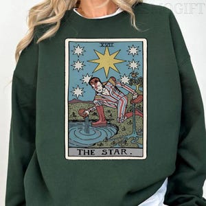 Puede incluir: Sudadera verde oscuro con un dise&ntilde;o de carta del tarot. La carta representa una figura vertiendo agua en una piscina, rodeada de estrellas, con el texto "THE STAR". El dise&ntilde;o es en tonos azules, rojos y dorados.