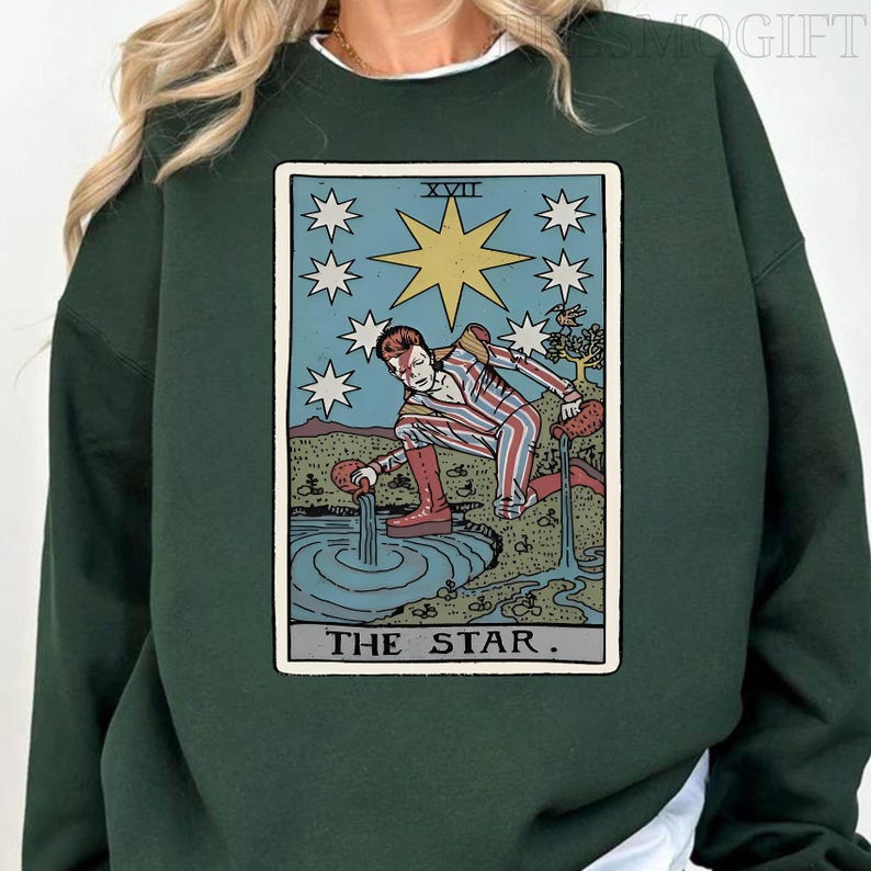 Puede incluir: Sudadera verde oscuro con un dise&ntilde;o de carta del tarot. La carta representa una figura vertiendo agua en una piscina, rodeada de estrellas, con el texto "THE STAR". El dise&ntilde;o es en tonos azules, rojos y dorados.