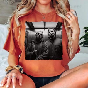 May include: Orange t-shirt featuring a black and white image of two horror movie characters in a lift. The characters are wearing masks and holding weapons.