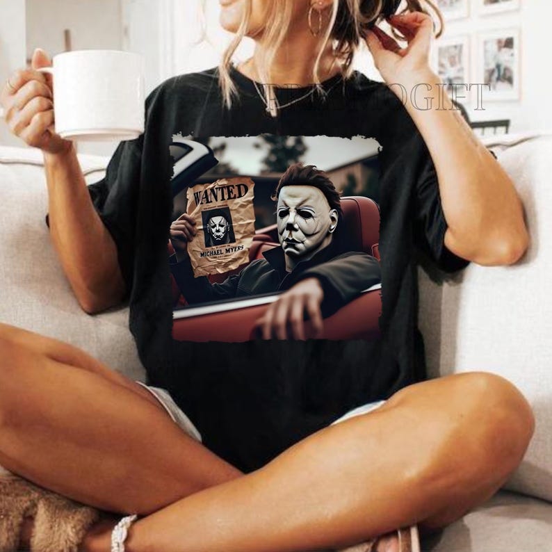May include: Black t-shirt featuring a graphic of Michael Myers from the Halloween films. The graphic depicts Michael Myers driving a car and holding a wanted poster with his own face on it. The poster reads "WANTED MICHAEL MYERS".