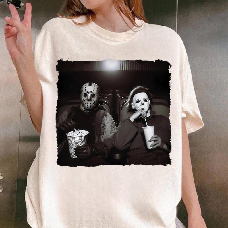 May include: A white t-shirt featuring a black and white image of two horror movie characters, Jason Voorhees and Michael Myers, seated in a cinema. The characters are holding snacks and drinks.