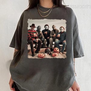 Op de afbeelding: Een donkergrijs T-shirt met een grafische print van horrorfilmfiguren die drankjes en pompoenen vasthouden. De personages zijn Freddy Krueger, Jason Voorhees, Michael Myers, Leatherface en Chucky.