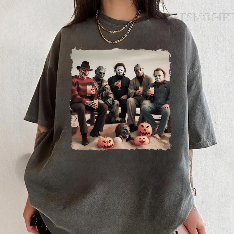 Op de afbeelding: Een donkergrijs T-shirt met een grafische print van horrorfilmfiguren die drankjes en pompoenen vasthouden. De personages zijn Freddy Krueger, Jason Voorhees, Michael Myers, Leatherface en Chucky.
