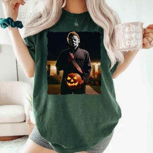 May include: A green t-shirt featuring a graphic print of a masked figure holding a jack-o'-lantern. The figure is standing in front of a house at night.