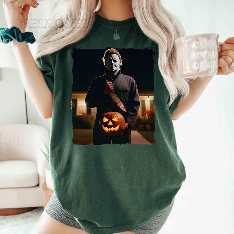May include: A green t-shirt featuring a graphic print of a masked figure holding a jack-o'-lantern. The figure is standing in front of a house at night.