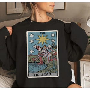 Puede incluir: Sudadera negra con dise&ntilde;o de carta del tarot. La carta representa una figura a rayas rojas y blancas vertiendo agua, rodeada de estrellas y una gran estrella amarilla. El texto "THE STAR" est&aacute; en la parte inferior.