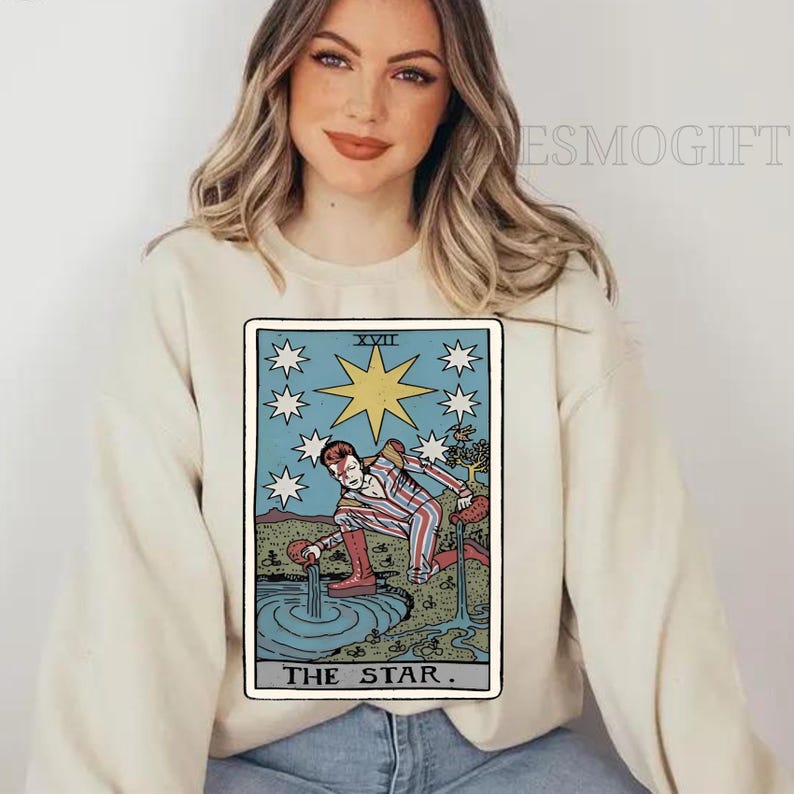 Puede incluir: Sudadera color crema con un dise&ntilde;o de carta del tarot. La carta representa una figura vertiendo agua en una piscina, rodeada de estrellas, con el texto "THE STAR". El dise&ntilde;o es en tonos azules, rojos y amarillos.