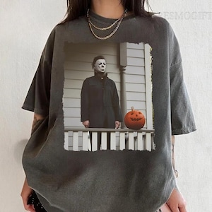 Op de afbeelding: Een donkergrijs T-shirt met een verweerde look met een geprinte afbeelding van een man met een wit masker en een zwart pak die op een veranda staat met een jack-o'-lantern.