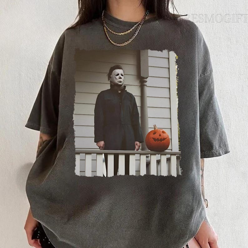 Op de afbeelding: Een donkergrijs T-shirt met een verweerde look met een geprinte afbeelding van een man met een wit masker en een zwart pak die op een veranda staat met een jack-o'-lantern.