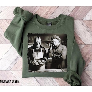 May include: A green sweatshirt with a black and white image of two horror movie characters in a kitchen. One character is holding a pan of cookies. The text "Military Green" is visible at the bottom of the image.