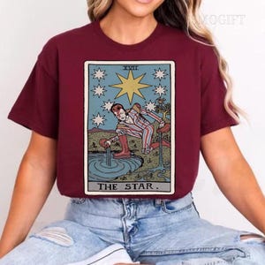 Puede incluir: Camiseta burdeos con un dise&ntilde;o de carta del tarot. El gr&aacute;fico muestra una figura vertiendo agua, rodeada de estrellas, con el texto "THE STAR". El dise&ntilde;o utiliza colores azul, beige y rojo.