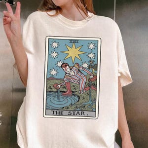 Puede incluir: Camiseta color crema con un dise&ntilde;o de carta del tarot. La carta representa una figura con ropa a rayas rojas y blancas, vertiendo agua de dos recipientes. La carta est&aacute; enmarcada con un borde azul y estrellas, con el texto "THE STAR."