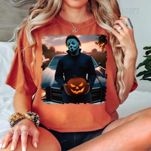 May include: An orange t-shirt featuring a graphic print of a man in a blue jumpsuit holding a jack-o'-lantern. The man is wearing a white mask and is standing in front of a vintage car.