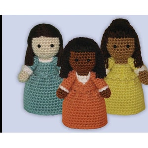 Puede incluir: Tres muñecas de crochet con diferentes tonos de piel y colores de cabello. Cada muñeca lleva un vestido. La muñeca de la izquierda lleva un vestido azul verdoso, la muñeca del centro lleva un vestido naranja y la muñeca de la derecha lleva un vestido amarillo.