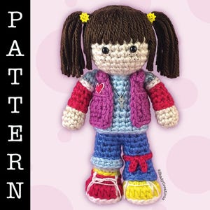 May include: Crocheted doll with brown hair, wearing a pink vest, blue shirt, blue jeans, and red and yellow shoes. The doll has a yellow smiley face on each pigtail.