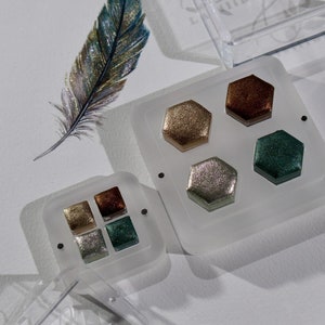 Könnte beinhalten: Zwei transparente, quadratische Lidschattenpaletten mit je vier sechseckigen Pfannen, gefüllt mit schimmerndem goldenem, braunem, silbernem und grünem Lidschatten. Eine dekorative Feder mit blauen, goldenen und silbernen Akzenten ist ebenfalls vorhanden.