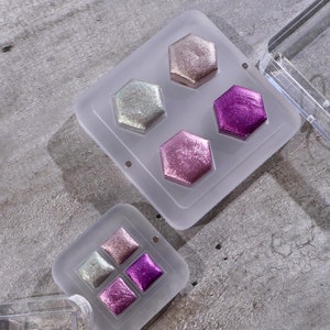 Könnte beinhalten: Zwei transparente Plastikpaletten mit sechseckigen und quadratischen Vertiefungen, gefüllt mit schimmernder Aquarellfarbe. Die Farben umfassen Rosa, Lila und Silber.