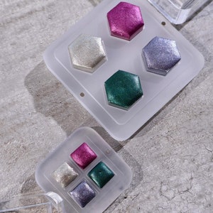 Könnte beinhalten: Ein Satz mit vier sechseckigen Farbtöpfen in einer durchsichtigen Plastikpalette. Die Farbtöpfe sind mit verschiedenen Farben gefüllt: rosa, silber, weiß und grün.