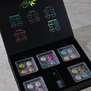 Könnte beinhalten: Eine schwarze Box mit mehreren kleinen, transparenten Kunststoffbehältern, die mit farbenfrohen, sechseckigen Artikeln gefüllt sind. Der Deckel der Box zeigt den Markennamen "Fisilinka" und verschiedene Produktnamen. Eine Flasche Nagellack ist ebenfalls enthalten.