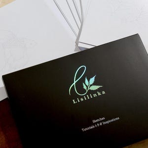 Könnte beinhalten: Eine schwarze Box mit dem Logo "Lisilinka" und dem Text "Sketches Tutorials 1-5 & Inspirations". Weiße Skizzenpapiere mit Fischzeichnungen sind sichtbar. Die Box hat einen holografischen Schimmer.