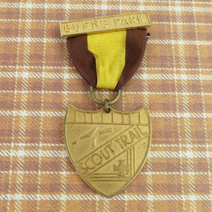 May include: A gold-toned metal medal featuring a shield-shaped pendant engraved with the text SCOUT TRAIL and a small knife icon. The pendant hangs from a yellow and brown fabric ribbon attached to a rectangular gold bar engraved with BURNS PARK.