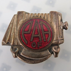 May include: Vintage metal pin with a camera design. The pin features a red circle with the letters "AA OF" in white. The pin has a bronze finish and is likely a collectible item.