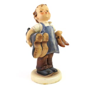 May include: A ceramic figurine of a young person holding two pairs of shoes. The figure wears a blue apron over a white shirt and brown pants. The shoes are brown and black, and the figurine is on a cream-colored base.