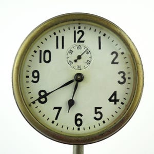 May include: A vintage clock with a white face, black numerals, and a smaller subdial. The clock has a gold-colored metal bezel and black hands. The time is approximately 7:40.
