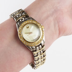 May include: A Movado watch with a silver and gold-tone band and bezel. The watch face is off-white with gold hands and a small gold dot at the 12 o'clock position. The bezel is adorned with small, clear crystals.