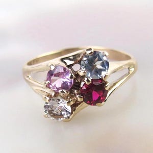 May include: A gold ring featuring four colorful gemstones: a light purple, a light blue, a deep red, and a clear white. The ring's band has a split design, with the gemstones clustered together.