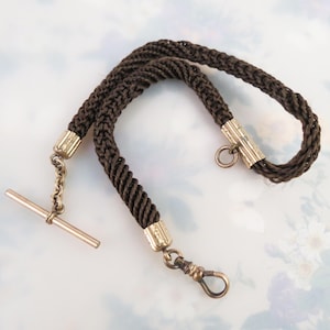 May include: A brown braided watch chain with gold-toned metal accents. The chain features a T-bar, a lobster clasp, and decorative metal caps. The braided design gives the chain a textured appearance, suitable for a vintage or antique aesthetic.