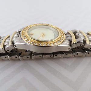 May include: A silver and gold-toned wristwatch with a cream-colored face. The watch features a gold bezel encrusted with small, clear crystals. The band is a combination of silver and gold links.
