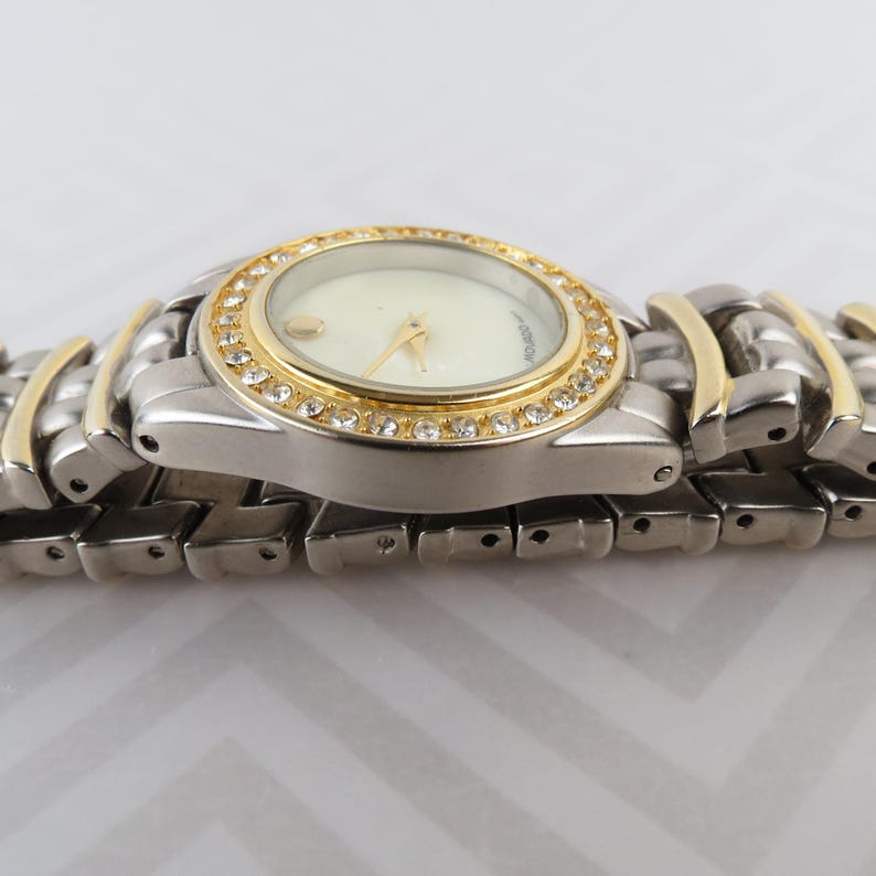 May include: A silver and gold-toned wristwatch with a cream-colored face. The watch features a gold bezel encrusted with small, clear crystals. The band is a combination of silver and gold links.