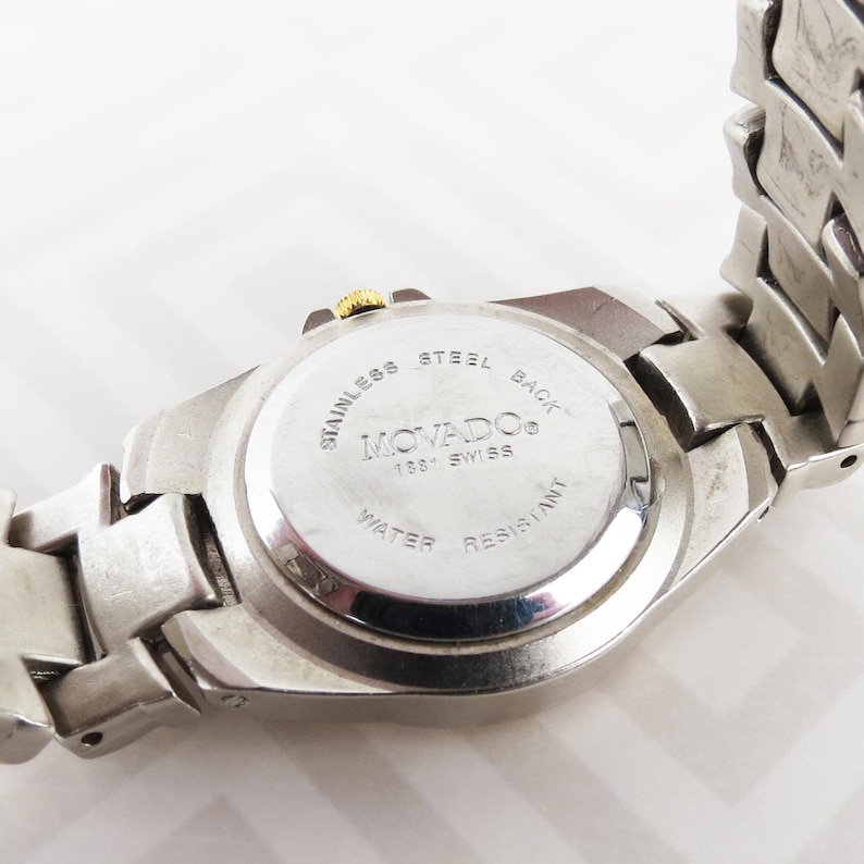 May include: A stainless steel Movado watch with a silver band. The watch face is visible, with the words "Stainless Steel Back" and "Water Resistant" printed on it. The watch has a gold-colored crown.