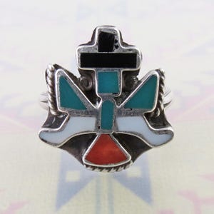 May include: A silver ring with a detailed inlaid design. The ring features a cross at the top and a stylized bird or angel shape below, with turquoise, black, white, and red accents. The ring has a textured rope-like border.