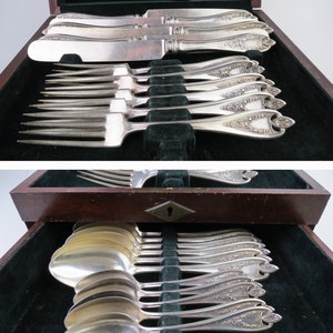 May include: A set of silver-plated flatware including knives, forks, and spoons, housed in a dark wooden storage chest. The handles feature an ornate floral pattern and a diamond-shaped tip. The interior lid displays the text 1847 ROGERS BROS.
