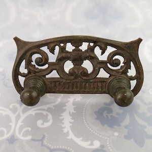 May include: Ornate, antique bronze-colored metal wall hook with a decorative openwork design. Features two rounded knobs and a scrolling, symmetrical pattern. Ideal for vintage or rustic decor.