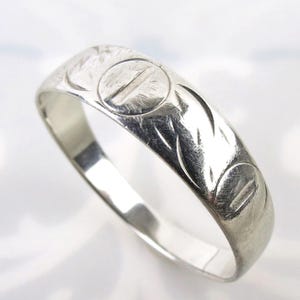 May include: A silver ring with engraved circular and curved leaf-like patterns. The ring has a slightly rectangular shape and a polished, reflective surface. The design is repeated around the band, creating a vintage or antique aesthetic.