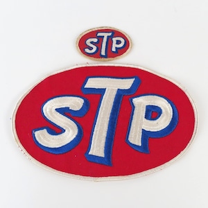 May include: Two STP patches. The larger patch is an oval shape with a red background and the letters "STP" in white with blue outlines. The smaller patch is also oval, with the same design. Both patches are embroidered.