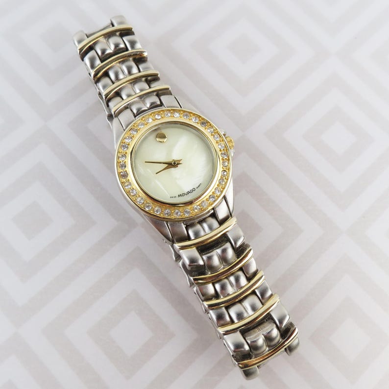 May include: A Movado wristwatch with a silver and gold-tone stainless steel band. The watch face is white with gold hands and a gold bezel encrusted with clear crystals. The watch is displayed on a white and gray patterned surface.