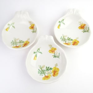 May include: Three white, shell-shaped ceramic dishes. Each dish features a yellow and green floral design. The flowers are a bright yellow with orange accents, and the leaves are green. The dishes are arranged on a white background.