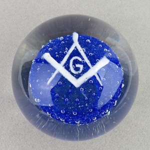 May include: A clear glass paperweight with a blue interior. The blue interior features a white Masonic symbol with the letter "G" in the center. The paperweight has small air bubbles throughout.