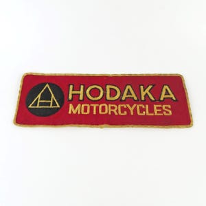 May include: A rectangular red patch with gold trim and lettering. The patch features the words "HODAKA MOTORCYCLES" in gold, with a black circle containing a gold triangle. The patch is likely for a motorcycle.