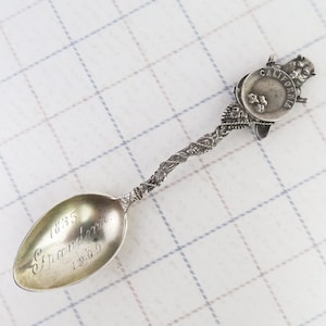 May include: Silver spoon with ornate handle and bowl. The bowl is engraved with the words "1835 Grandam 1909". The handle features a detailed floral design and a small charm with the word "CALIFORNIA" and a bear.