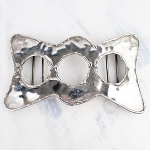 May include: A silver-toned metal belt buckle with a hammered, textured surface. The buckle features an elongated, bow-like shape with three circular cutouts arranged horizontally, each containing a thin metal bar for securing a belt strap.