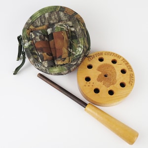 May include: A wooden turkey call with the text "Clifton Custom Turkey Calls" and a turkey head design. Includes a camouflage carrying case and a wooden striker. The call has multiple holes and a light wood finish.