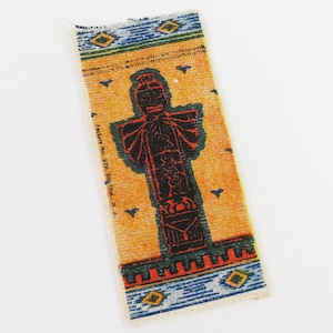 May include: Rectangular textile with a central black figure on an orange background. The figure has a detailed design with red and green accents. The textile has blue and yellow geometric patterns on the top and bottom edges. The text "Factory No. 279. 5th Dist. N.J." is printed on the side.