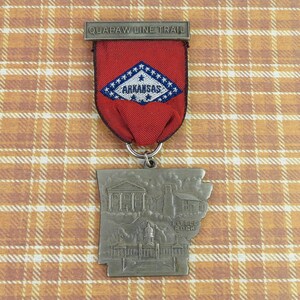 May include: A metal souvenir medal featuring a red fabric ribbon with a diamond-shaped patch displaying the text ARKANSAS and QUAPAW LINE TRAIL. The medal is shaped like the state of Arkansas and depicts embossed buildings and the text LITTLE ROCK.