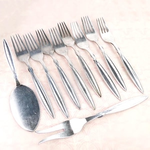 May include: A collection of stainless steel silverware, including forks and a serving spoon, arranged on a light pink patterned surface. The forks have a slender, curved handle design. The image showcases a set of dining utensils.